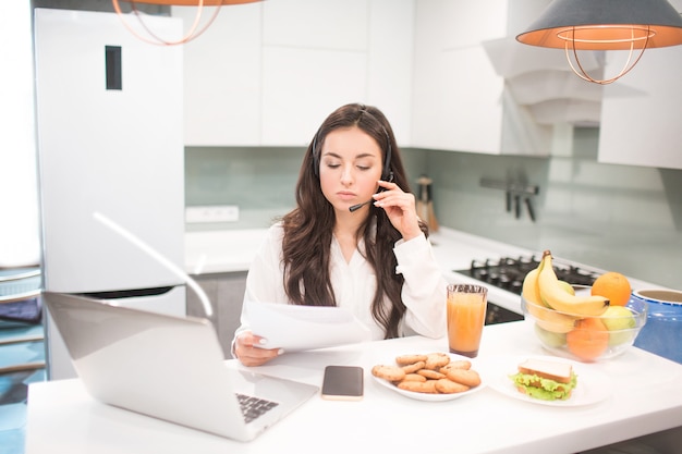 Hermosa mujer de cabello negro trabaja desde casa y usa auriculares con auriculares. Un empleado se sienta en la cocina y tiene mucho trabajo en una computadora portátil y tableta y tiene videoconferencias y reuniones.