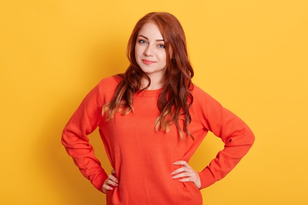 Hermosa mujer con cabello ondulado rojo, mirando directamente a la cámara mientras mantiene las manos en las caderas, posando en un suéter casual naranja sobre fondo amarillo.