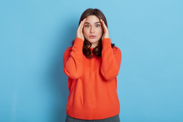 Hermosa mujer con cabello oscuro, mantiene las manos cerca de los ojos, viste un jersey naranja casual, posando aislada en la pared azul, mira con ojos grandes a la cámara, manteniendo el gesto de escondite.