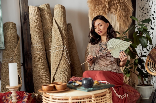 Hermosa mujer de cabello oscuro en traje étnico chamánico en habitación decorada al estilo de bali está haciendo ceremonia de rappa