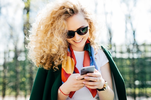 Hermosa mujer con cabello rizado con gafas de sol y chaqueta verde con smartphone en sus manos