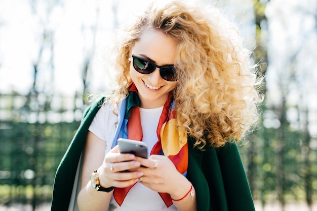 Hermosa mujer con cabello rizado con gafas de sol y chaqueta verde sosteniendo un teléfono inteligente en sus manos navegando por Internet disfrutando de la comunicación en línea mientras está de pie contra el fondo verde del parque