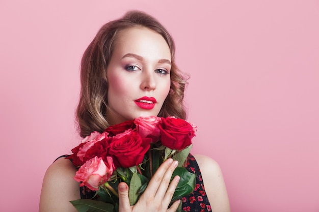 Hermosa mujer con cabello rizado y labios rojos con ramo de rosas sobre fondo rosa