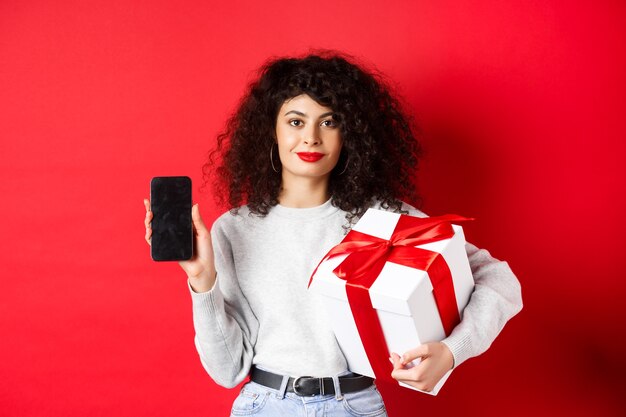 Hermosa mujer con cabello rizado, mostrando la aplicación de compras en la pantalla vacía del teléfono inteligente, sosteniendo el regalo envuelto en una caja festiva, de pie en la pared roja.