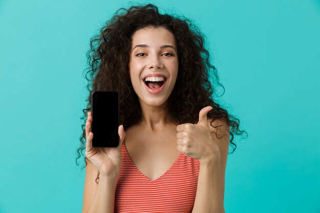 Hermosa mujer con cabello rizado sonriendo y sosteniendo un teléfono celular, aislado sobre una pared azul