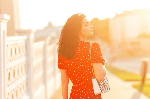 Hermosa mujer con cabello rizado en un vestido rojo vintage con bolsos de moda camina en la ciudad al atardecer