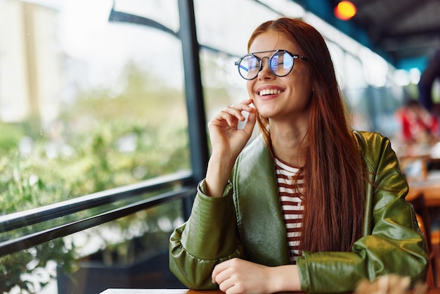 Foto hermosa mujer con cabello rojo y anteojos se sienta en un café en la ciudad y mira por la ventana sonriendo con dientes bloguera independiente femenina closeup