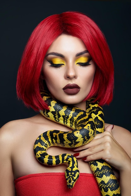 Hermosa mujer con cabello rojo y una serpiente pitón amarilla alrededor de su cuello. maquillaje de ojos de belleza brillante profesional lápiz labial amarillo, rojo burdeos