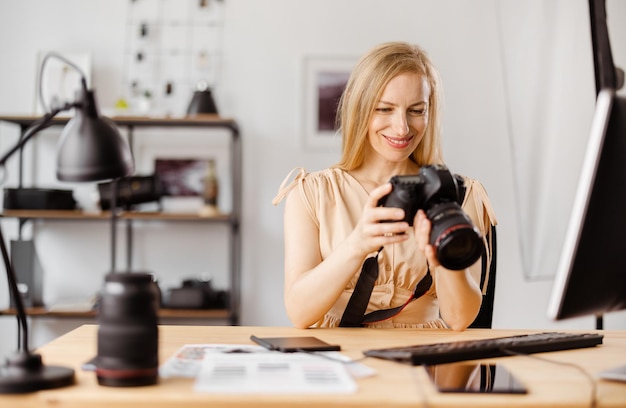 Hermosa mujer con cabello rubio sentado en la oficina y revisando fotos en la cámara Fotógrafa eligiendo fotos para editarlas en la computadora