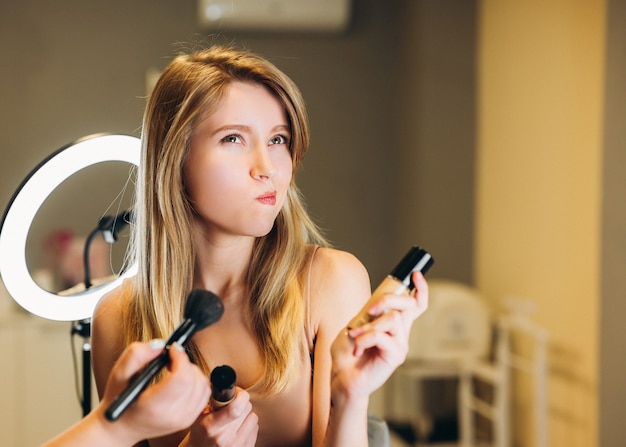 Hermosa mujer con cabello rubio tiene cosméticos en sus manos. Una clienta maquillada no está contenta con el resultado.