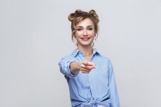 La hermosa mujer, con camisa azul, con miradas de felicidad, señalando con el dedo a la cámara. Disparo de estudio aislado sobre fondo gris