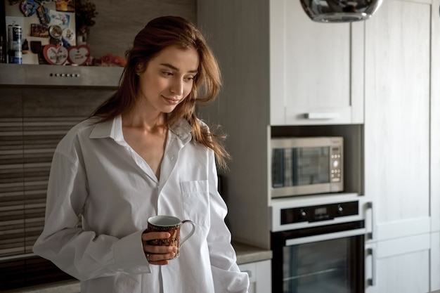 Hermosa mujer en camisa blanca, sentado con una taza de té en la cocina y hablando por teléfono móvil