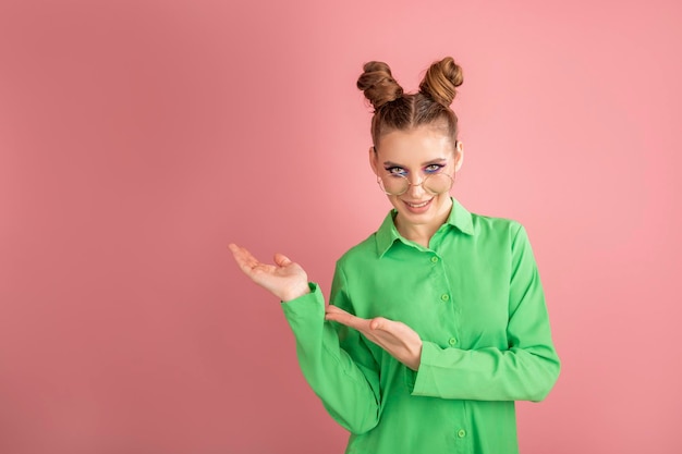 Hermosa mujer con camisa verde sobre fondo rosa, mostrando y señalando algo que está en su mano. Copie el espacio.