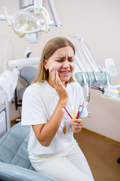 Una hermosa mujer con una camiseta blanca se sienta en un consultorio dental, se mira en el espejo, sonríe y sostiene un cepillo de dientes en sus manos. Chica con aparatos ortopédicos muestra cuidado bucal. Odontología, tratamiento dental