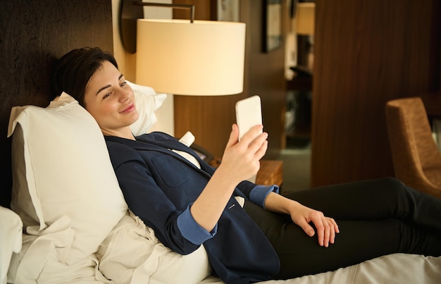 Foto hermosa mujer cansada en traje de negocios, sonríe tiernamente mirando a cámara, usando un teléfono móvil mientras está acostado en la cama en el dormitorio