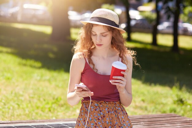 Hermosa mujer cargando su teléfono en el cargador de panel solar incorporado en un banco sentado en el parque de la ciudad