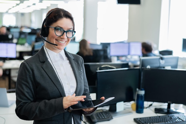 Foto hermosa mujer caucásica con auriculares sostiene una tableta digital mientras está de pie en una oficina de espacio abierto