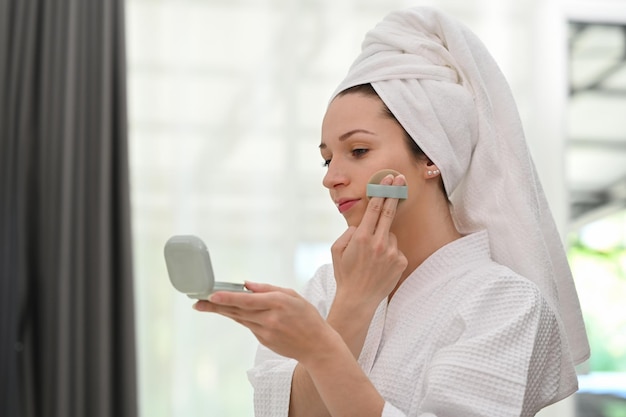 Hermosa mujer caucásica en bata de baño con cojín y espejo aplicando base de maquillaje en polvo en la cara