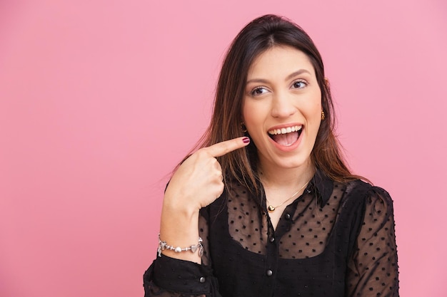 Foto hermosa mujer caucásica brasileña fondo rosa apuntando a la mejilla y sonrisa concepto de apariencia de belleza estética