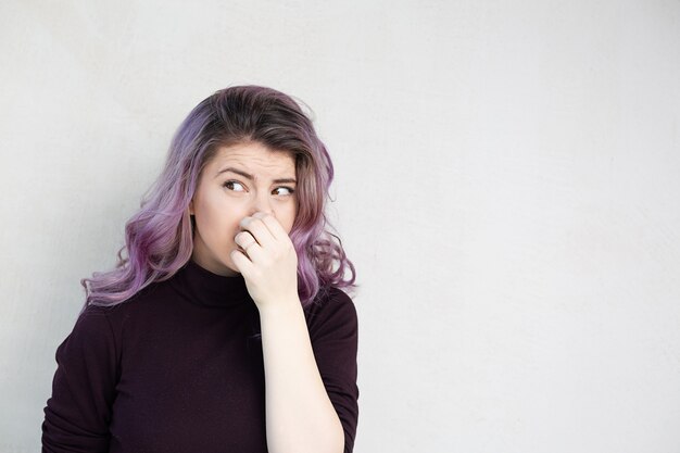 Hermosa mujer caucásica con cabello morado conteniendo la respiración con los dedos en la nariz. Concepto de olores repugnantes. Espacio para texto