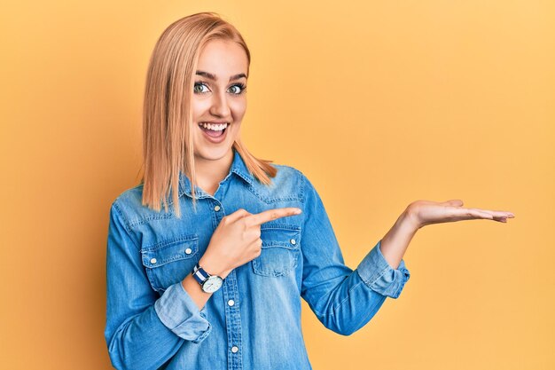 Hermosa mujer caucásica con una chaqueta de denim casual sorprendida y sonriendo a la cámara mientras presentaba con la mano y señalaba con el dedo.