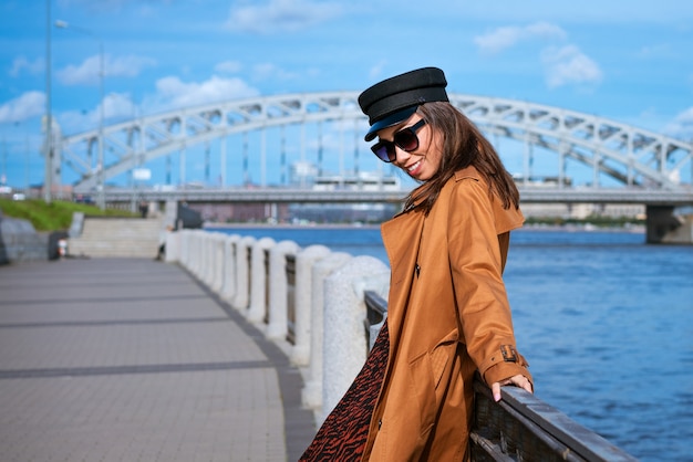 Hermosa mujer caucásica en gafas de sol, con una gorra negra y chaqueta posando mientras está de pie en el terraplén en un día soleado contra una pared de cielo azul y paisaje urbano