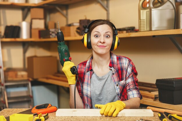 Hermosa mujer caucásica joven de cabello castaño en camisa a cuadros, auriculares con aislamiento acústico que trabajan en el taller de carpintería en el lugar de la mesa, perforando con taladros eléctricos en la pieza de madera, haciendo muebles.