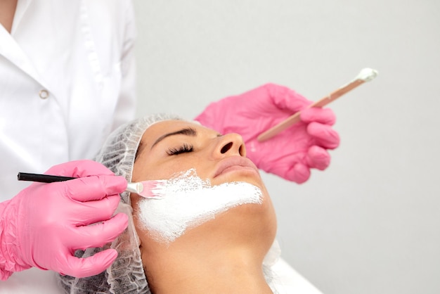 Hermosa mujer caucásica joven satisfecha utiliza los servicios de esteticista profesional en salón de belleza o spa Esteticista irreconocible aplicó máscara blanca a la mitad de la cara del cliente con cepillo Vista superior