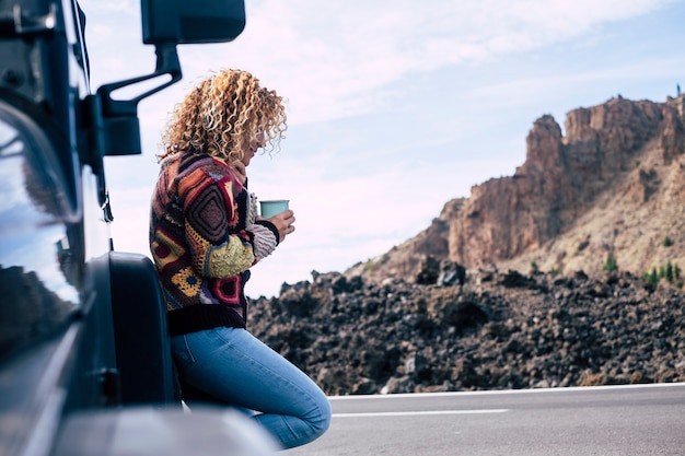 Hermosa mujer caucásica de mediana edad rubia rizada independiente descansar después de un viaje con su automóvil negro todoterreno estacionado cerca de la calle.