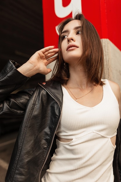 Hermosa mujer caucásica de moda con peinado en elegante chaqueta de cuero negro y poses de vestido blanco en la ciudad en la calle