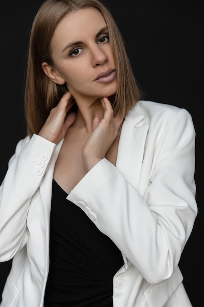 Hermosa mujer caucásica de moda con un vestido negro y una chaqueta blanca en el estudio con un fondo negro mirando a la cámara