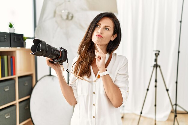 Hermosa mujer caucásica que trabaja como fotógrafa en un estudio de fotografía pensando concentrada en la duda con el dedo en la barbilla y mirando hacia arriba preguntándose