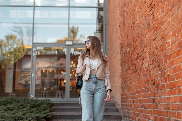Hermosa mujer caucásica en ropa casual de moda con gafas de sol, chaqueta de cuero de moda con jeans vintage de cintura alta y una bolsa caminando en la ciudad cerca de un centro comercial