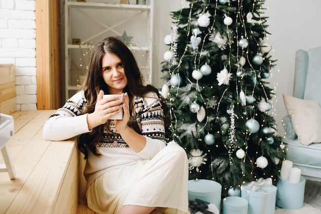Hermosa mujer caucásica sonriente con taza sentada cerca del árbol de Navidad decorado con estilo