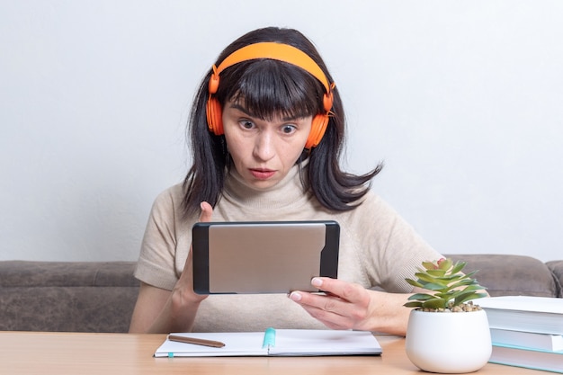 Hermosa mujer caucásica sorprendida en auriculares naranja usando una tableta mientras está sentado en la mesa en casa