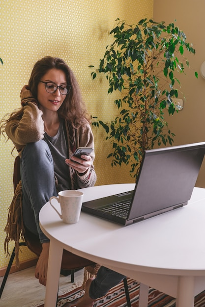 Hermosa mujer caucásica con teléfono móvil y sentado en la mesa con laptop y taza de café