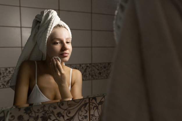 Hermosa mujer caucásica con una toalla en la cabeza en el baño después de una ducha se mira en el espejo