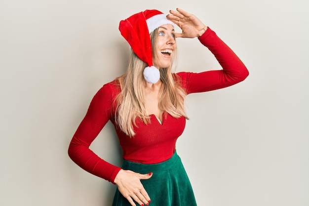 Hermosa mujer caucásica con traje de navidad y sombrero muy feliz y sonriente mirando lejos con la mano sobre la cabeza. concepto de búsqueda.