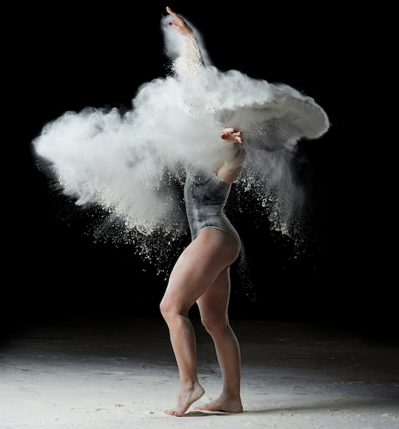 Hermosa mujer caucásica en un traje negro con una figura deportiva está bailando en una nube blanca de harina