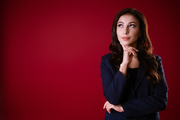 Hermosa mujer caucásica en un traje sobre un fondo rojo con una mirada pensativa toca su barbilla con el ...