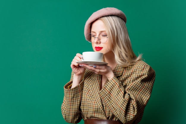 Hermosa mujer en chaqueta de estilo y sombrero con taza en pared verde