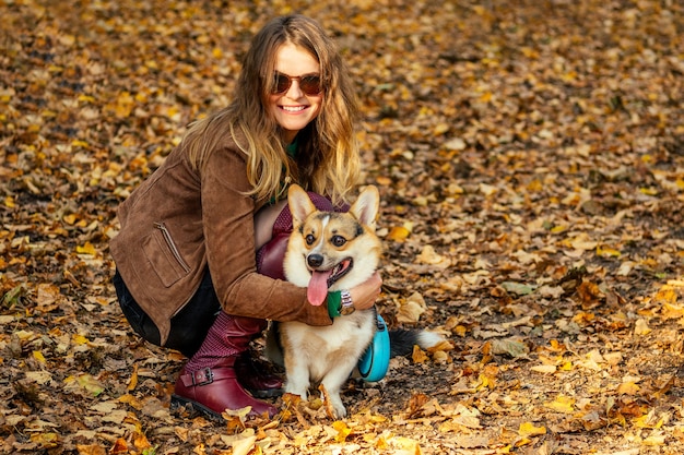 Hermosa mujer con una chaqueta marrón y elegantes botas rojas sentada sobre hojas amarillas caídas junto a su mascota corgi y abrazando a un cachorro de otoño bonito día.La ternura y el amor de la dueña de un perro