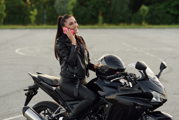 Hermosa mujer ciclista habla por teléfono inteligente mientras está sentado en una elegante motocicleta deportiva en el estacionamiento al aire libre