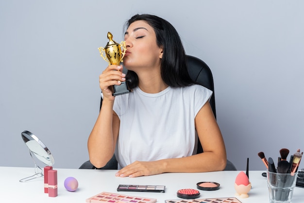 Hermosa mujer complacida se sienta a la mesa con herramientas de maquillaje sosteniendo y besando la copa ganadora