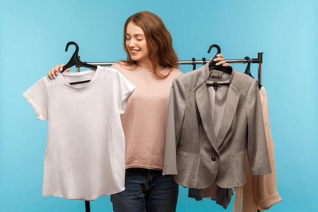 Hermosa mujer compradora positiva sosteniendo ropa en bastidores y mirando la camisa eligiendo ropa de estilo casual en lugar de compras clásicas de chaqueta de negocios en una boutique moderna en un estudio interior aislado