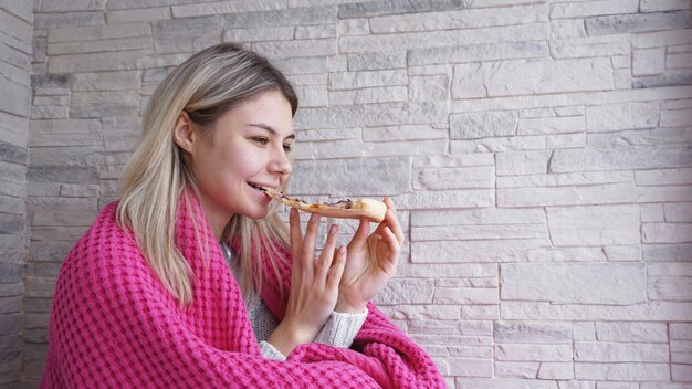 Hermosa mujer con cuadros rosas sostiene una rebanada de pizza en la mano y se la come. De cerca