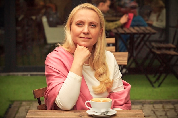 Una hermosa mujer de cuarenta años vestida con una cicatriz rosa está tomando café en un café.