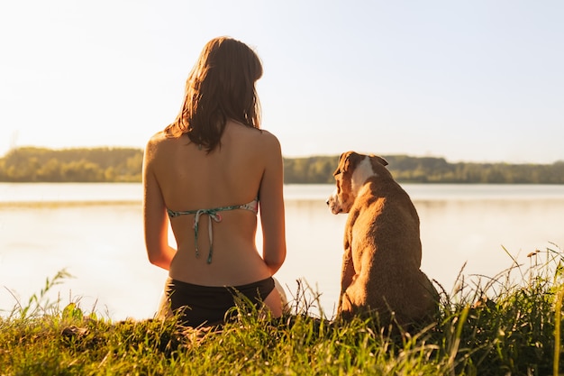 Hermosa mujer delgada con perro mascota disfrutando de hermosa vista cerca del lago al atardecer de la cálida tarde soleada. Young fit hembra sentada con perro staffordshire terrier en la orilla del río