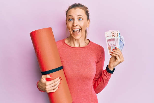 Hermosa mujer deportiva caucásica sosteniendo colchoneta de yoga y billetes de coronas suecas celebrando loco y asombrado por el éxito con los ojos abiertos gritando emocionado.