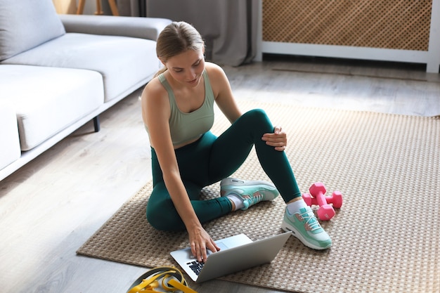 Hermosa mujer deportiva delgada en ropa deportiva está sentada en el suelo con pesas y está usando una computadora portátil en casa en la sala de estar. Estilo de vida saludable. Quédese en las actividades de la casa.